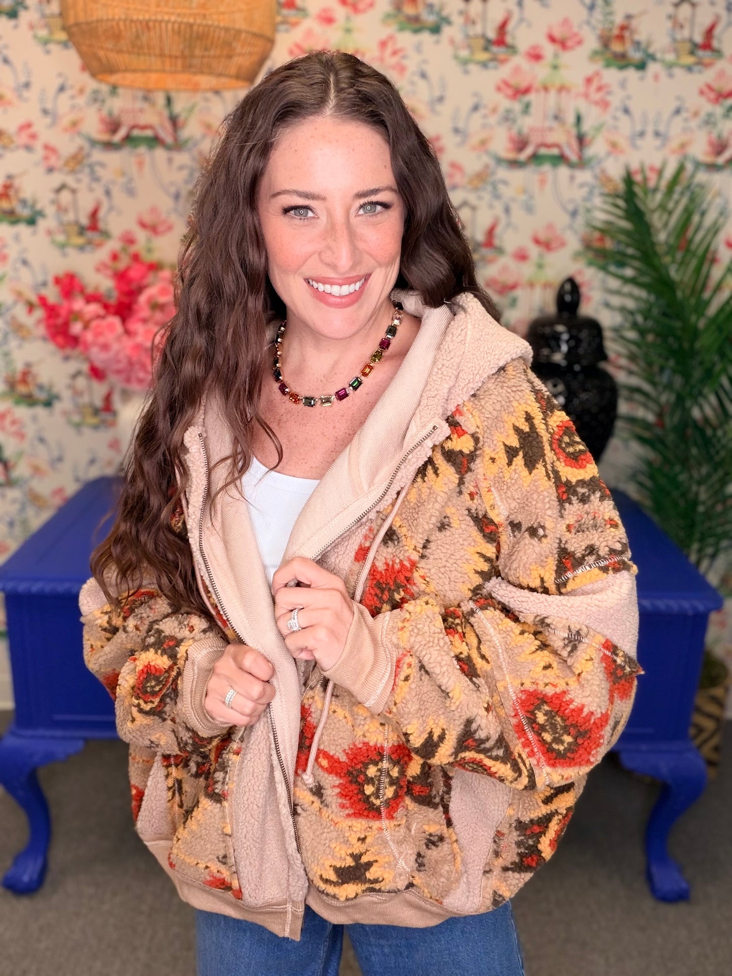 A woman smiling while wearing a beige sherpa hoodie with a colorful floral patterned outer layer, standing in front of a floral wallpaper background with a blue bench.