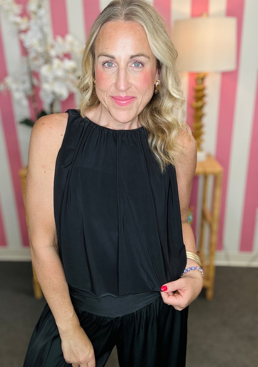 A woman wearing a sleeveless black top with a gathered, pleated bodice and a matching black skirt, standing in front of a pink and white striped wall with a lamp in the background.