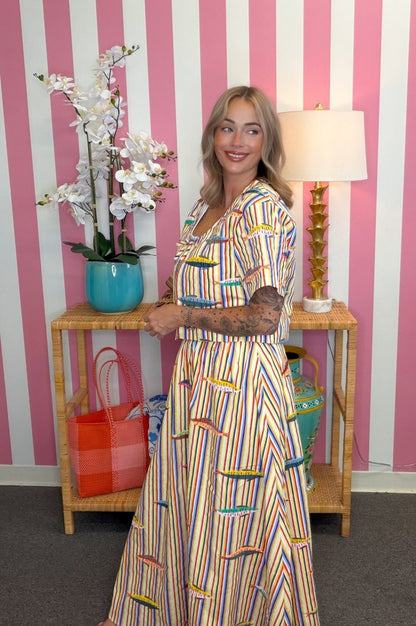 Woman in a colorful dress standing in front of a striped pink and white wall with a table and lamp.