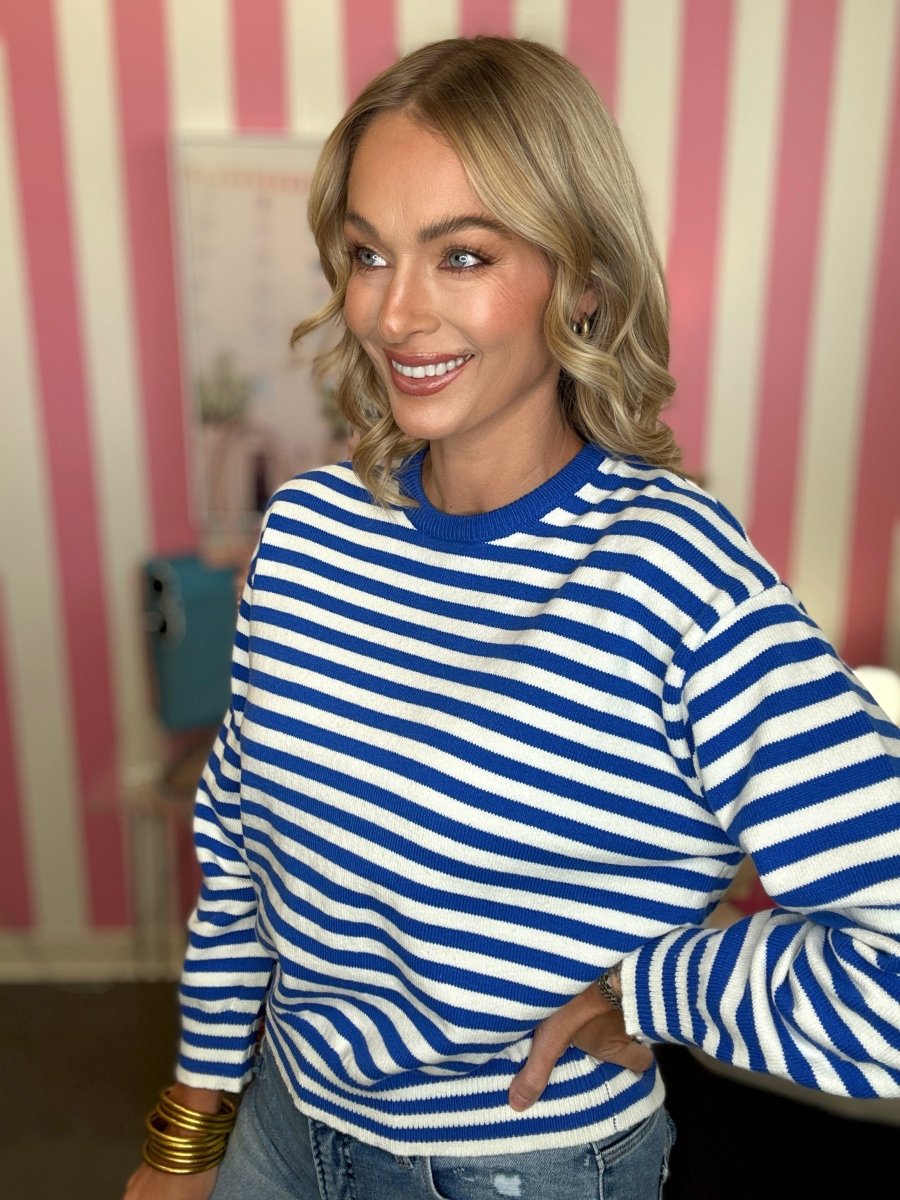 A woman with blonde wavy hair smiling and looking to the side, wearing a blue and white horizontally striped long-sleeve knit top with a solid blue crew neck, paired with blue jeans. She is standing in front of a pink and white vertically striped wall.