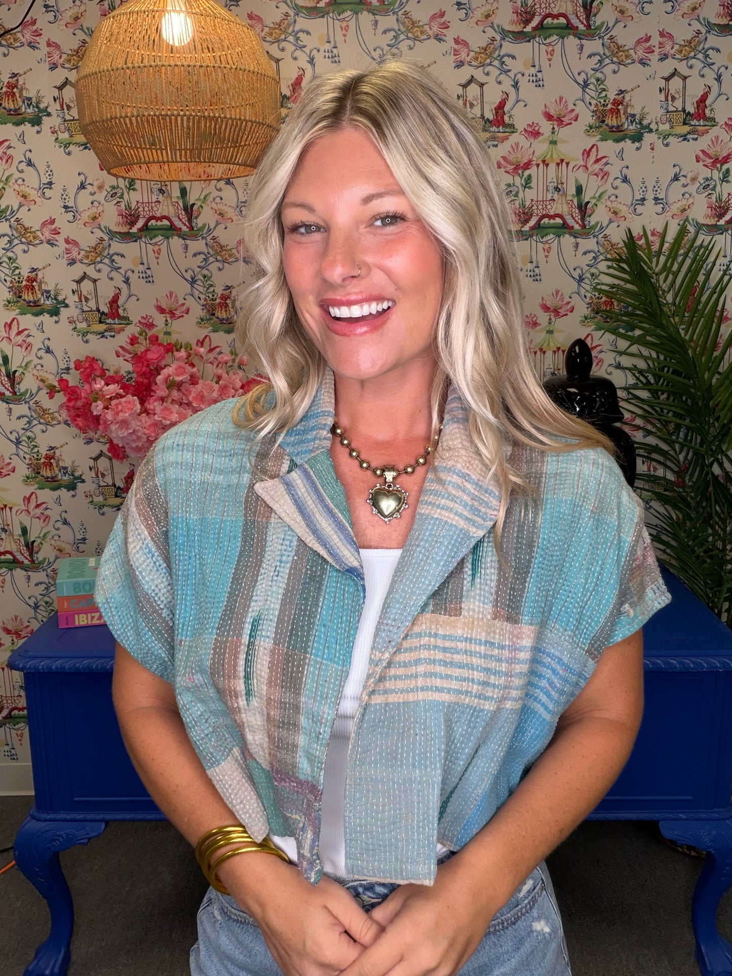 A smiling blonde woman wearing a light blue and beige striped short-sleeve button-up shirt over a white top, standing in front of a floral wallpaper background with a blue table and woven pendant light.