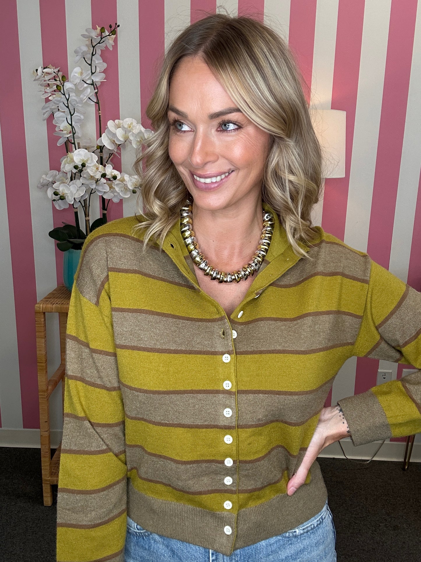 A woman wearing a yellow-green and brown striped button-up cardigan sweater, styled with a chunky beaded necklace and blue jeans, standing in front of a pink and white striped wall.