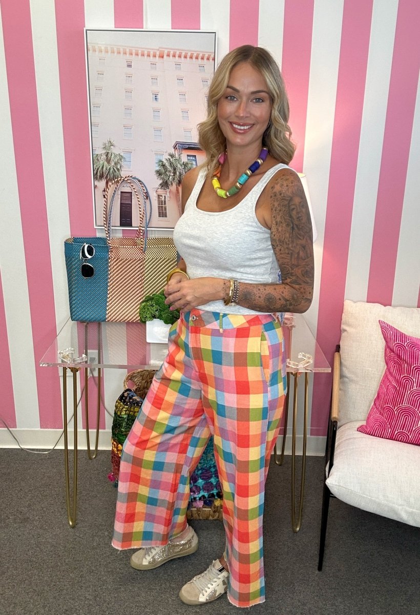 A woman stands in a room with pink and white striped walls, wearing a white tank top and colorful plaid wide-leg pants. She is accessorized with a chunky rainbow beaded necklace, bracelets, and metallic sneakers.