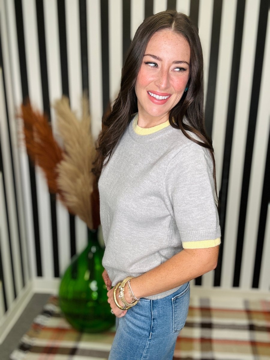 A woman wearing a light gray crewneck sweater with yellow ribbed collar and sleeve cuffs, paired with blue jeans, standing in front of a black and white striped wall.