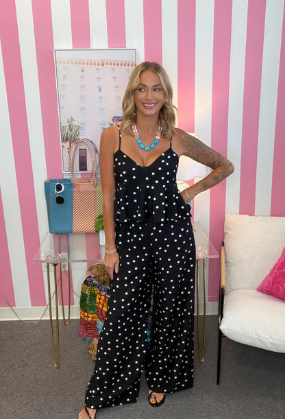 A woman stands indoors wearing a black sleeveless jumpsuit with white polka dots. The room has pink and white vertical striped walls, a small glass table with a blue bag, and a white chair with a pink pillow.