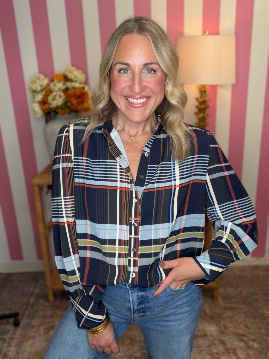 A smiling blonde woman wearing a long-sleeve button-up plaid shirt with navy, blue, white, orange, and yellow stripes, paired with blue jeans, posed in front of a pink and white striped wall.