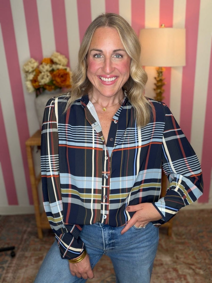 A smiling blonde woman wearing a long-sleeve button-up plaid shirt with navy, blue, white, orange, and yellow stripes, paired with blue jeans, posed in front of a pink and white striped wall.