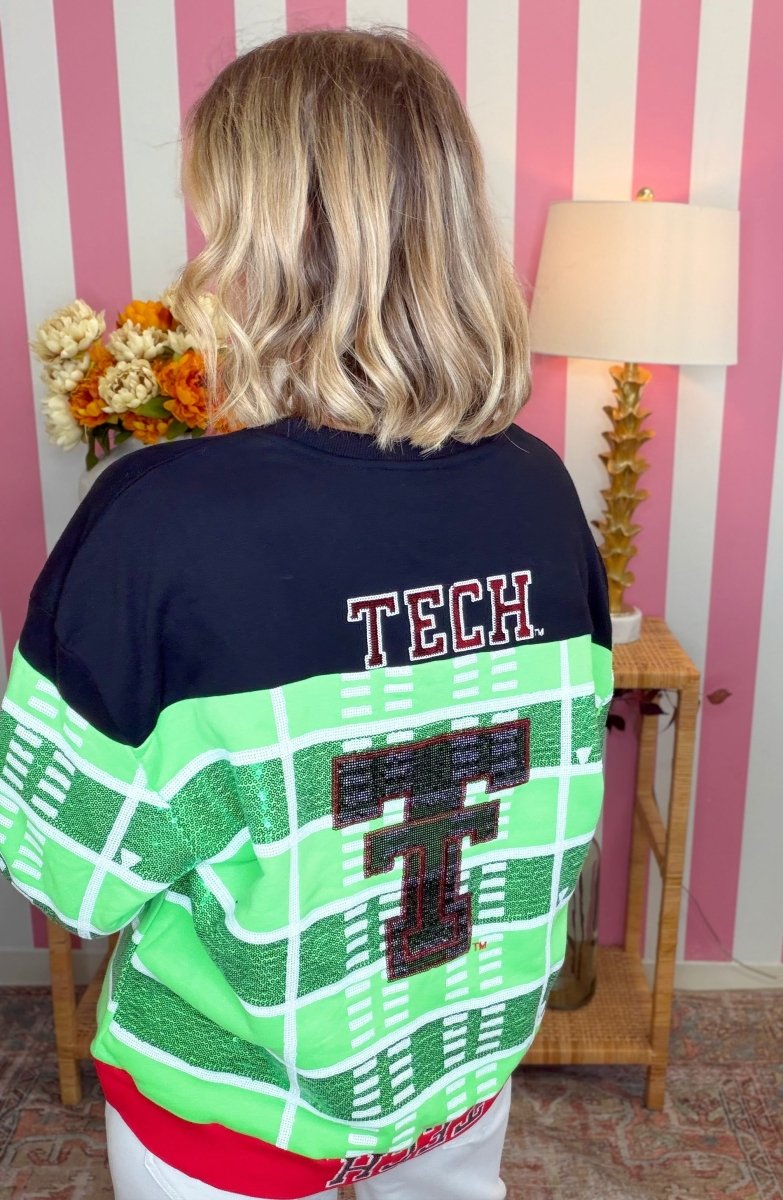 A person viewed from behind wearing a short-sleeve athletic-style sweatshirt with a navy upper section, bright green lower section, white grid lines, and large 'T' graphics on the back. The room has pink-and-white striped wallpaper, a lamp, flowers, and a side table.