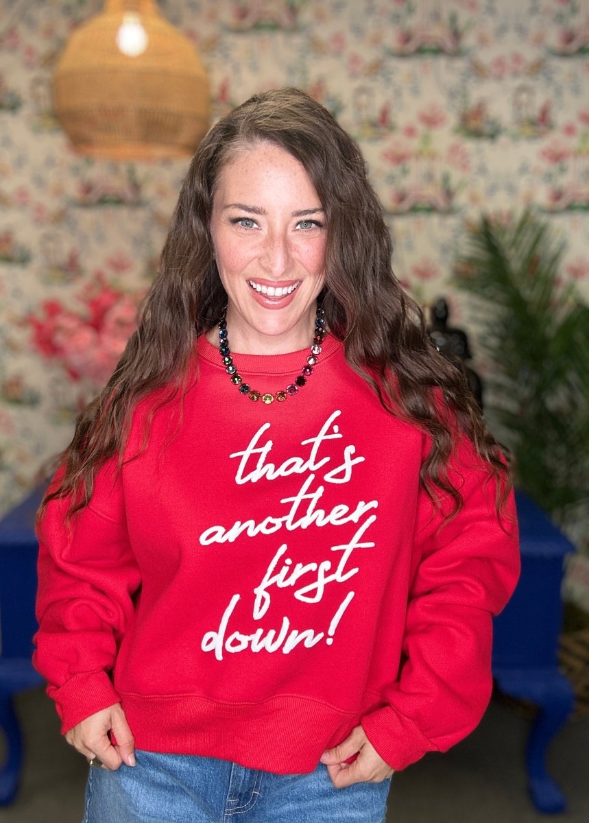 A woman smiling while wearing a red crewneck sweatshirt with the phrase 'that's another first down!' printed in white script across the front, paired with blue jeans in a cozy, rustic indoor setting.