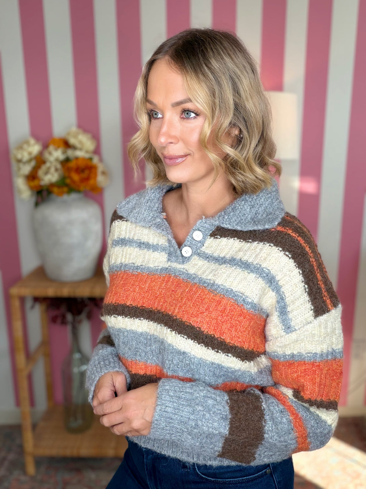 A woman wearing a fuzzy knit polo-style sweater with horizontal stripes in light blue, cream, orange, and brown, standing in front of pink and white striped wallpaper.