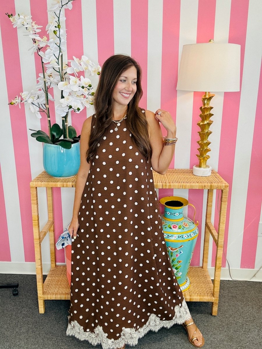 Woman in a brown polka dot dress standing in front of a pink and white striped wall with decorative vases and a lamp.