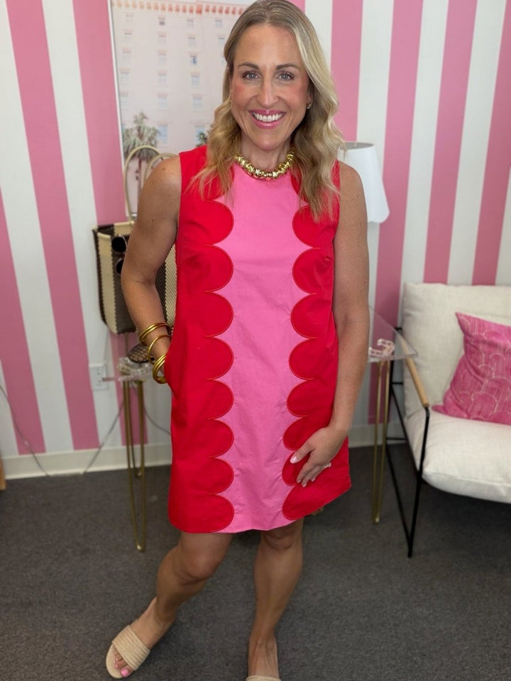 A woman modeling a sleeveless dress with a bold pink and red color-blocked design featuring scalloped ruffle details down the front, posed in front of a pink and white striped wall.