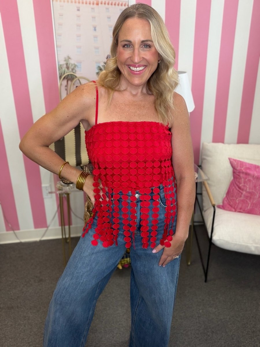 A woman wearing a vibrant red sleeveless top with a textured circular pattern and dangling pom-pom fringe, paired with blue jeans, standing in front of a pink and white striped wall.