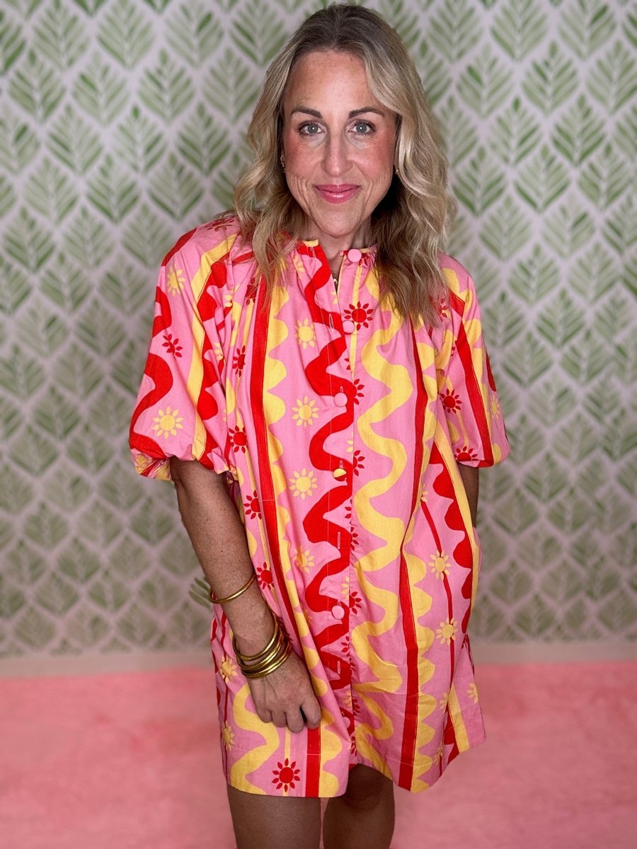 A woman wearing a short-sleeve pink button-up dress with a bold red and yellow abstract pattern, standing against a green patterned wall and pink floor.