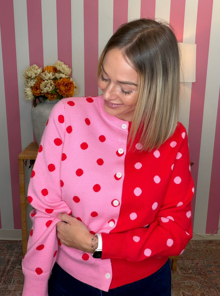 A woman wearing a split-color cardigan sweater with one half in pink and the other in red, both featuring white polka dots. She is standing in front of a pink and white striped wall.