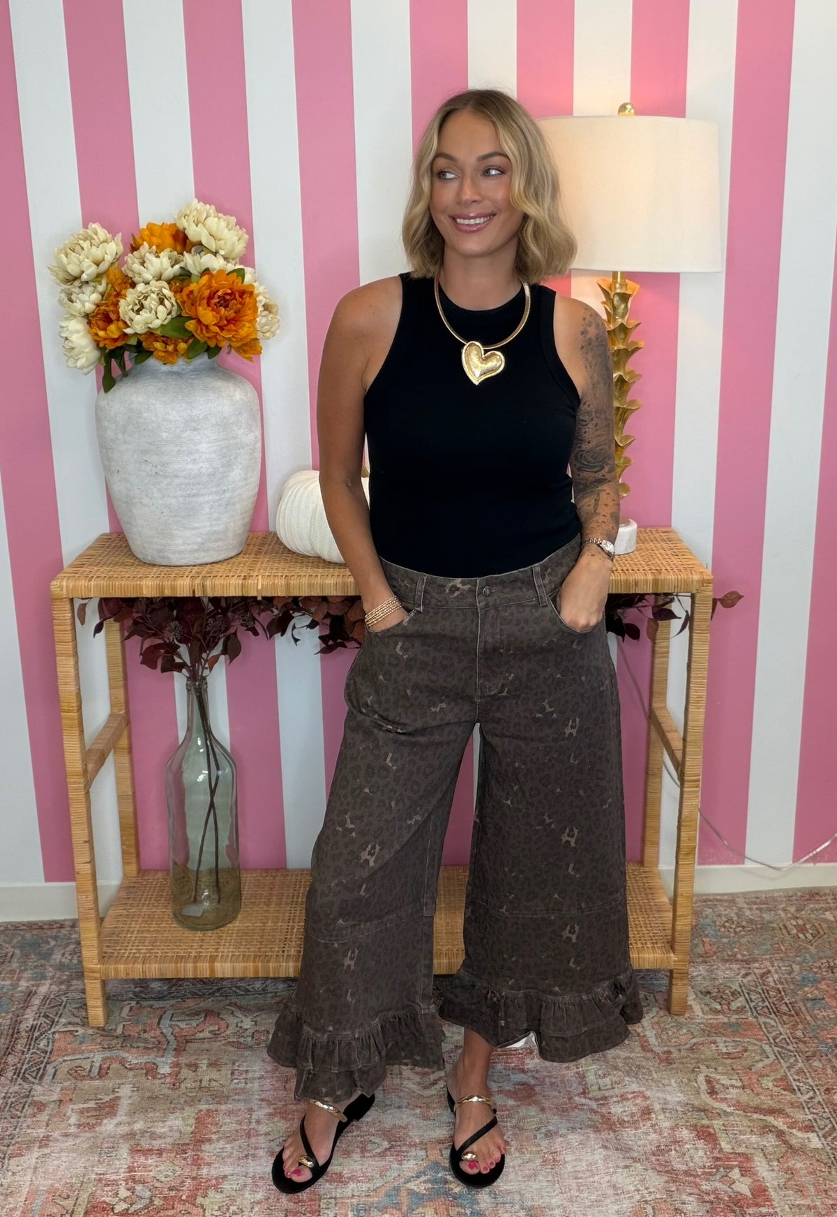 A woman stands in a room with pink and white striped wallpaper, wearing a black sleeveless top and brown wide-leg ruffled pants. She is standing in front of a wooden console table decorated with a vase of flowers and a lamp.