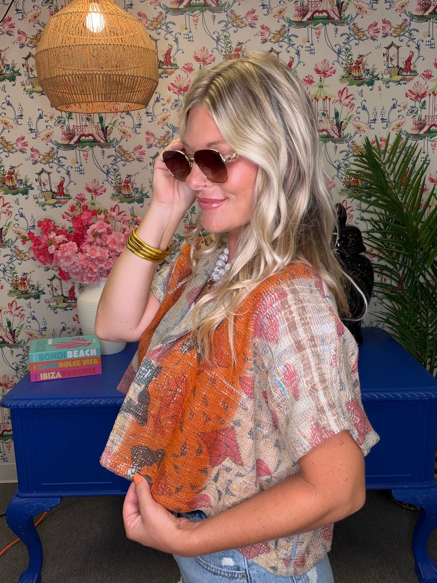 A woman modeling a brown and beige plaid apron over a light blue and white plaid shirt, standing in a decorated room with floral wallpaper, a blue side table, and a wicker pendant light.