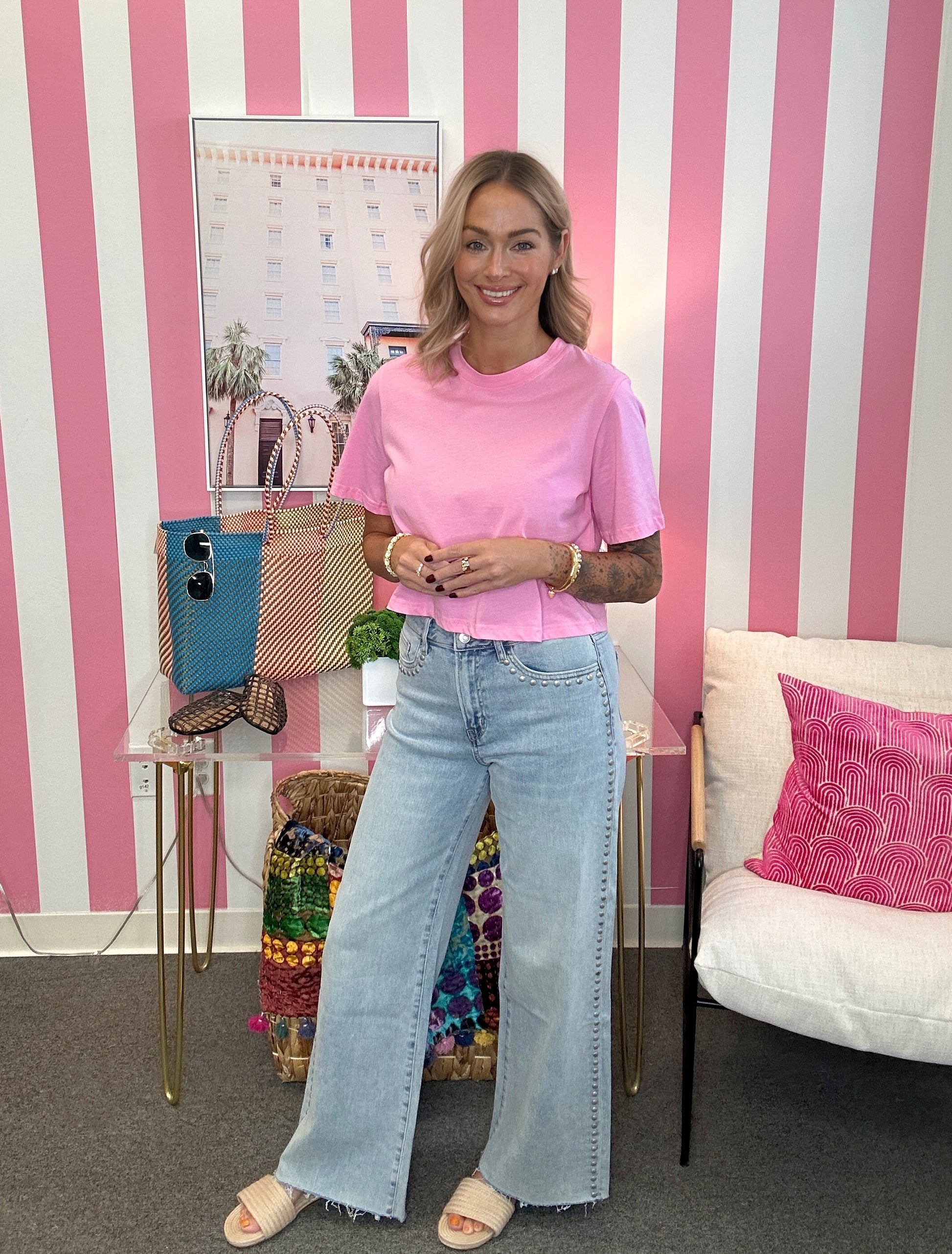 A woman stands in a room with pink and white striped wallpaper, wearing a light pink short-sleeve t-shirt and light-wash wide-leg jeans. She is standing next to a glass side table with a blue bag and a white chair with a pink pillow.