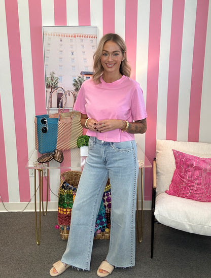 A woman stands in a room with pink and white striped wallpaper, wearing a light pink short-sleeve t-shirt and light-wash wide-leg jeans. She is standing next to a glass side table with a blue bag and a white chair with a pink pillow.