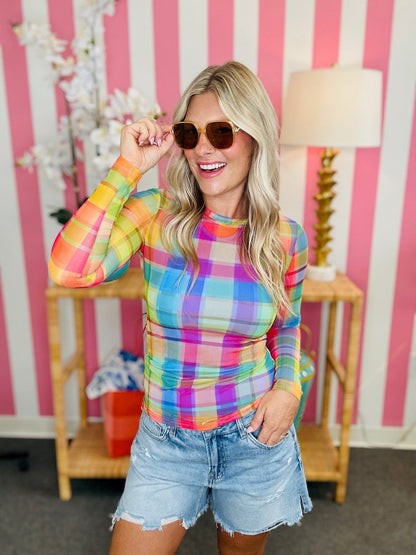 Woman wearing a colorful plaid shirt and denim shorts in front of a pink and white striped wall.