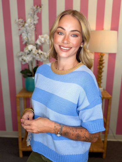 A smiling blonde woman wearing a light blue and navy striped knit sweater with a beige crew neck, standing in front of a pink and white striped wall with a lamp and side table in the background.