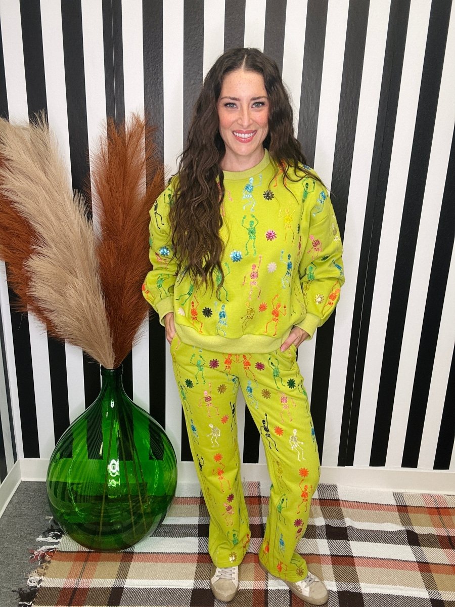 A woman stands indoors wearing a bright yellow-green matching lounge set with colorful embroidered details. She is posed in front of a black-and-white striped wall, with a large green glass vase and pampas grass to her left and a plaid rug beneath her feet.