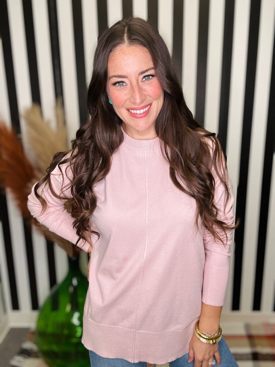 A smiling woman with long wavy brown hair wearing a light pink mock-neck long-sleeve top and blue jeans, standing in front of a black and white striped background.