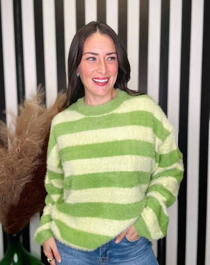 A woman smiling while wearing a fuzzy, long-sleeve knit sweater with horizontal lime green and cream stripes, paired with blue jeans, set against a black and white striped background.