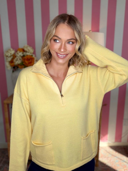 A woman wearing a pale yellow quarter-zip pullover sweater with a mock neck and front pockets, posing in front of a pink and white striped wall.
