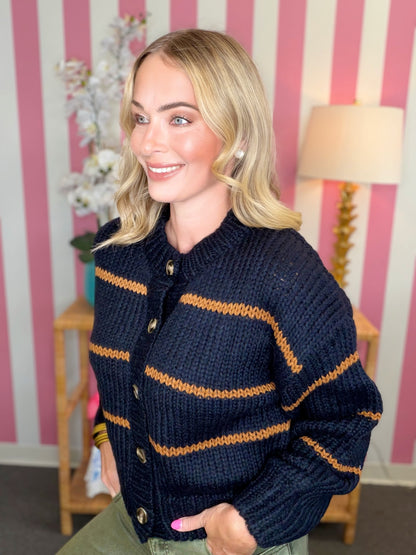 A smiling blonde woman wearing a chunky knit navy blue cardigan with horizontal mustard yellow stripes, posed in front of a pink and white striped wall.