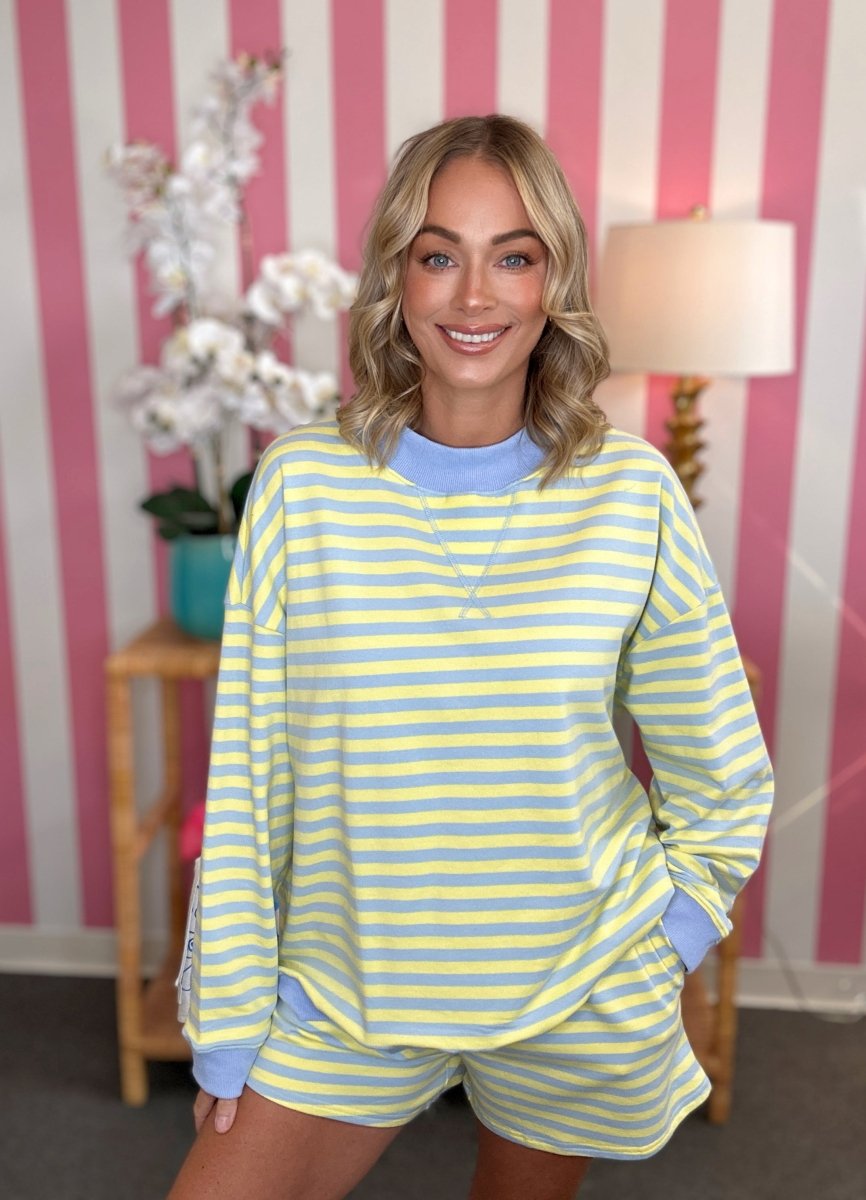 A woman modeling a matching yellow and light blue striped lounge set, featuring a long-sleeve top and shorts, posed in front of a pink and white striped wall.