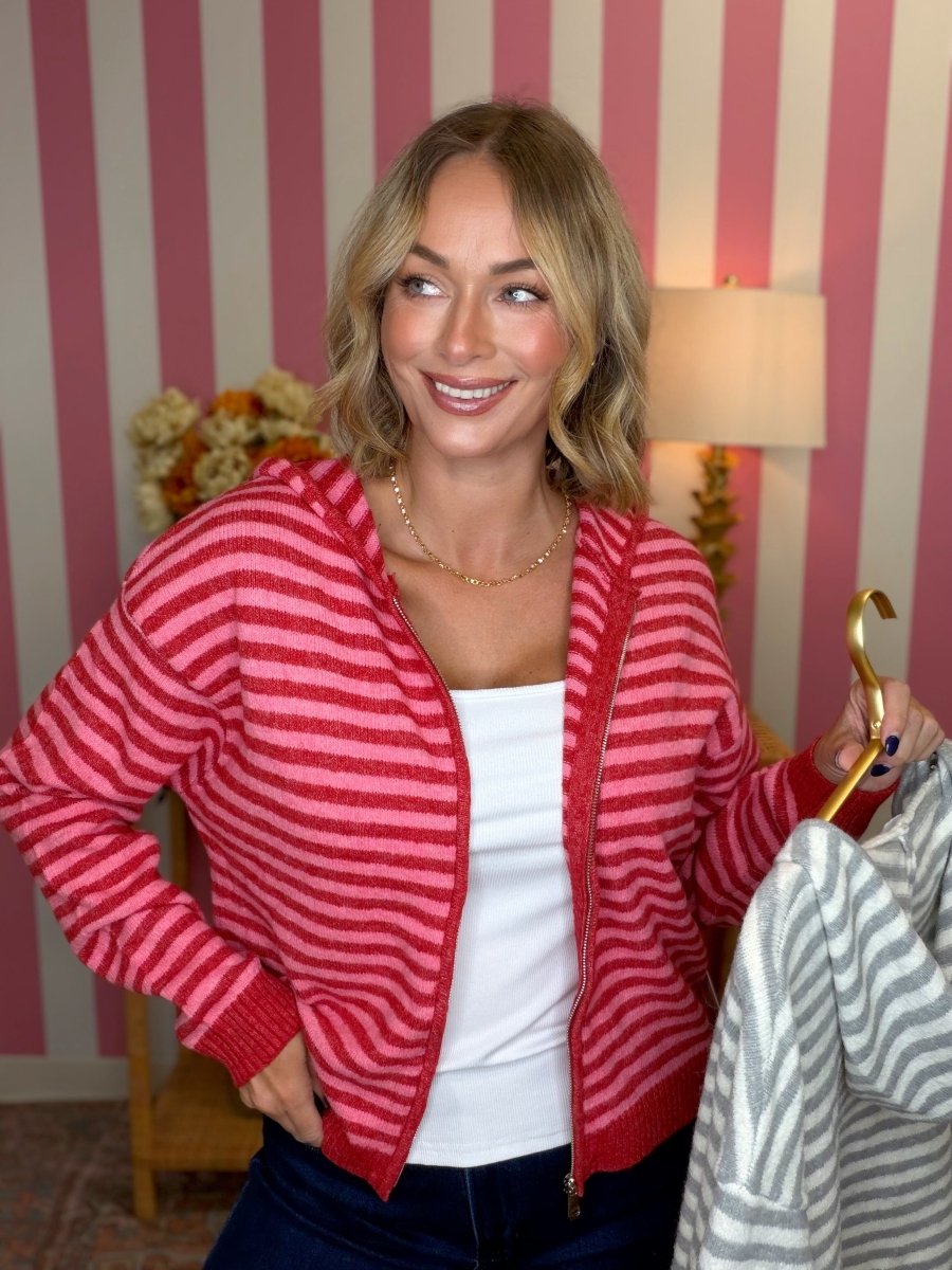 woman standing in a pink and red striped hoodie holding a grey and white stiped hoodie on a gold hanger