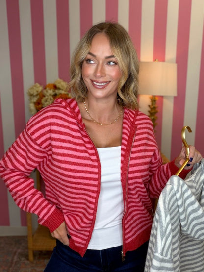 woman standing in a pink and red striped hoodie holding a grey and white stiped hoodie on a gold hanger