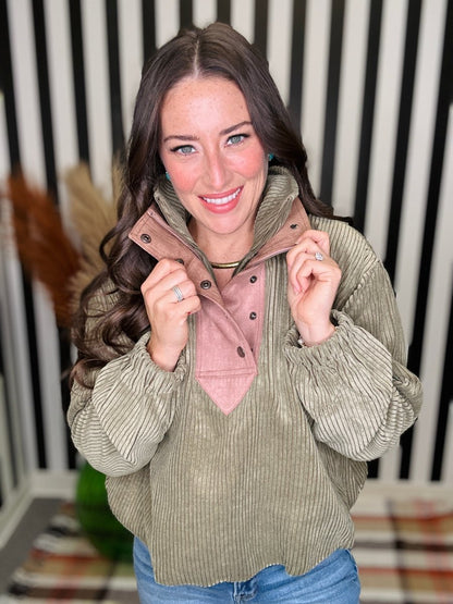 A woman wearing a sage green corduroy pullover with a pink mock neck collar, posing in front of a black and white striped background.