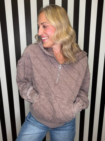 A woman wearing a taupe-brown diamond-quilted half-zip pullover hoodie with a drawstring hood and front pockets, paired with blue jeans, standing in front of a black and white striped wall.