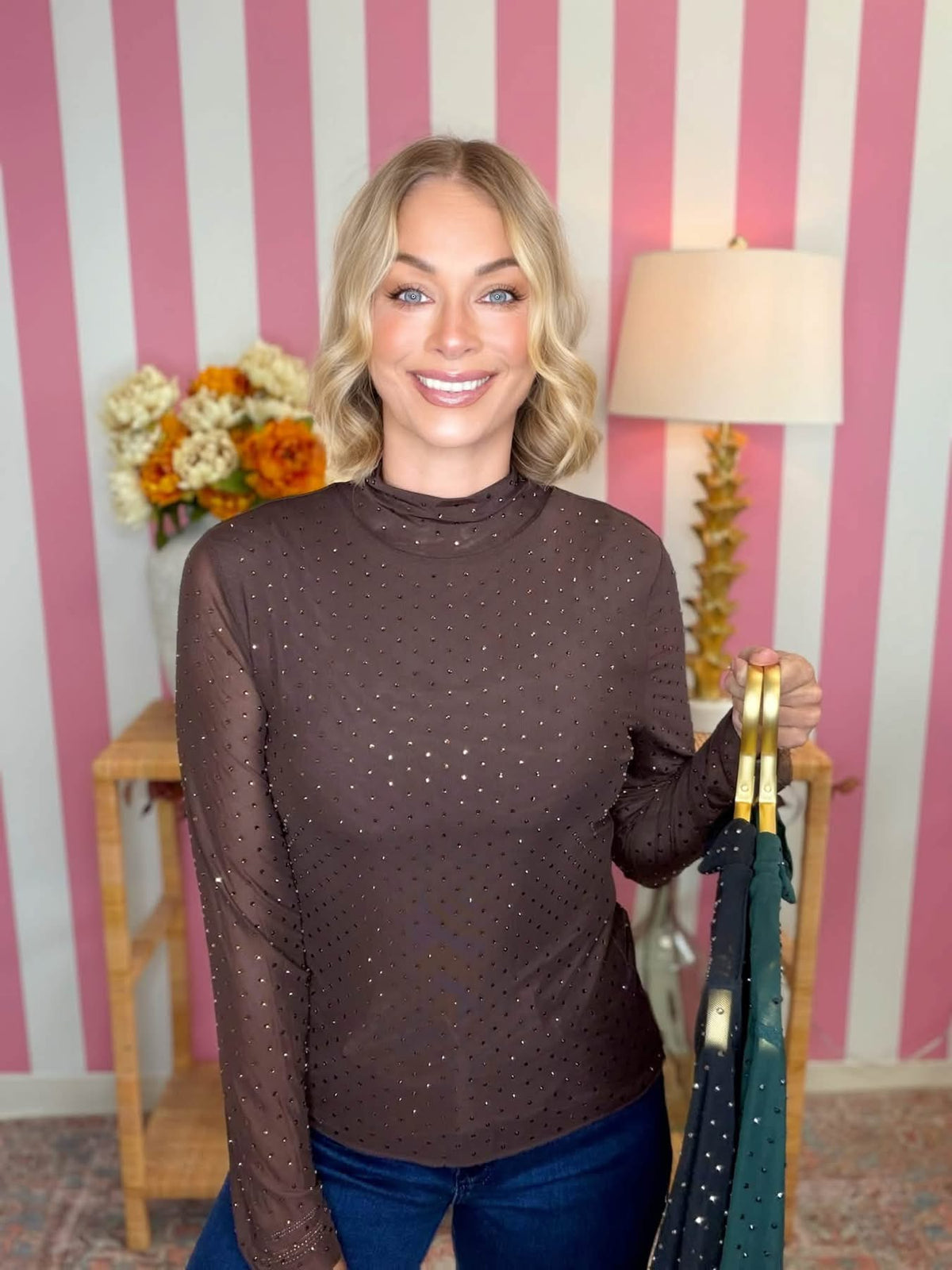 A smiling blonde woman wearing a dark brown sheer long-sleeve mock neck top with subtle glittery dots, paired with blue jeans. She is holding up a dark green sheer top with gold hangers in front of a pink and white striped wall background.