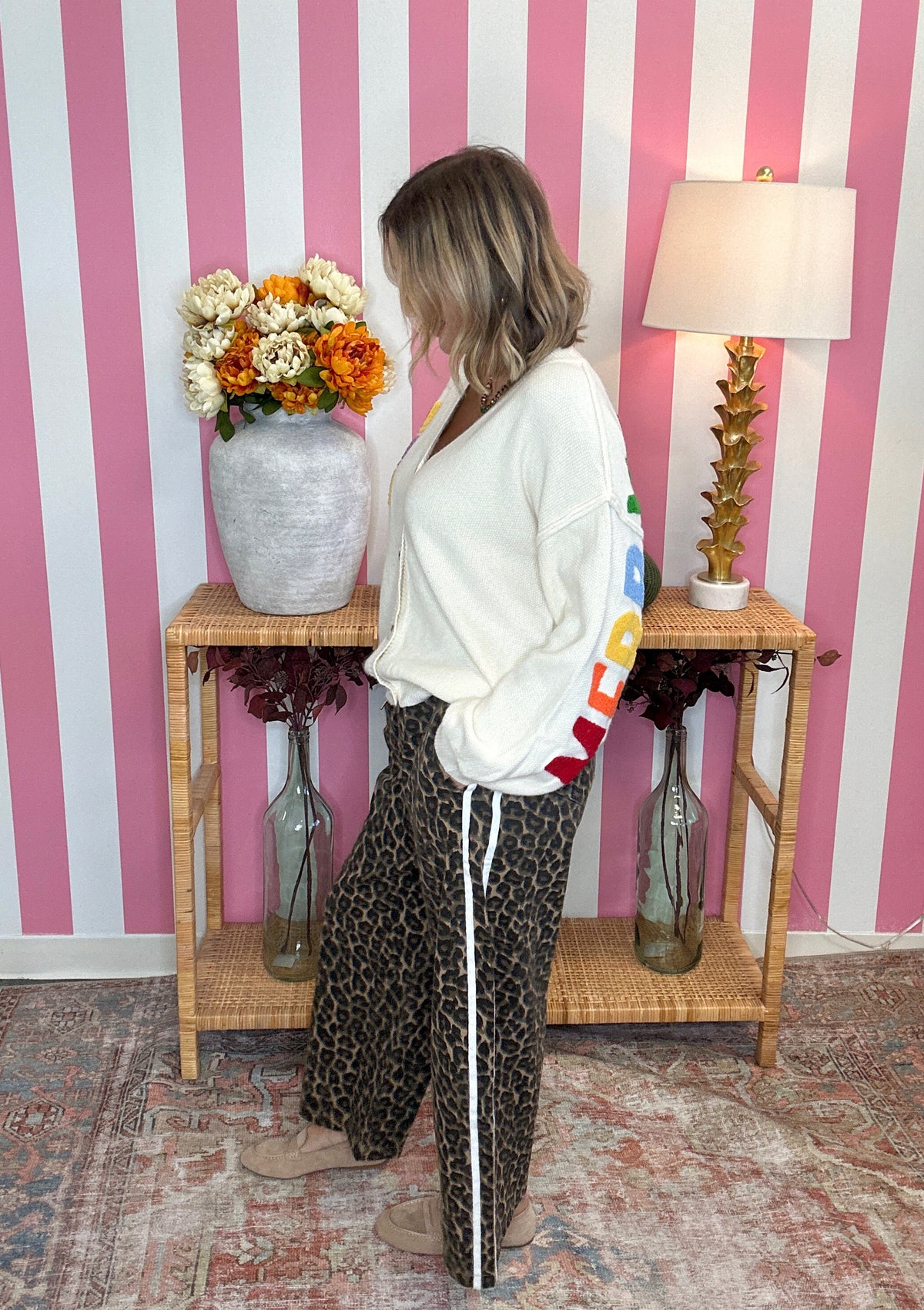 A woman stands in profile wearing a white cropped sweatshirt with a colorful graphic print on the sleeve and dark leopard-print pants with a white side stripe. She is posed in front of a pink and white striped wall with a wooden side table holding a vase of flowers and a lamp.