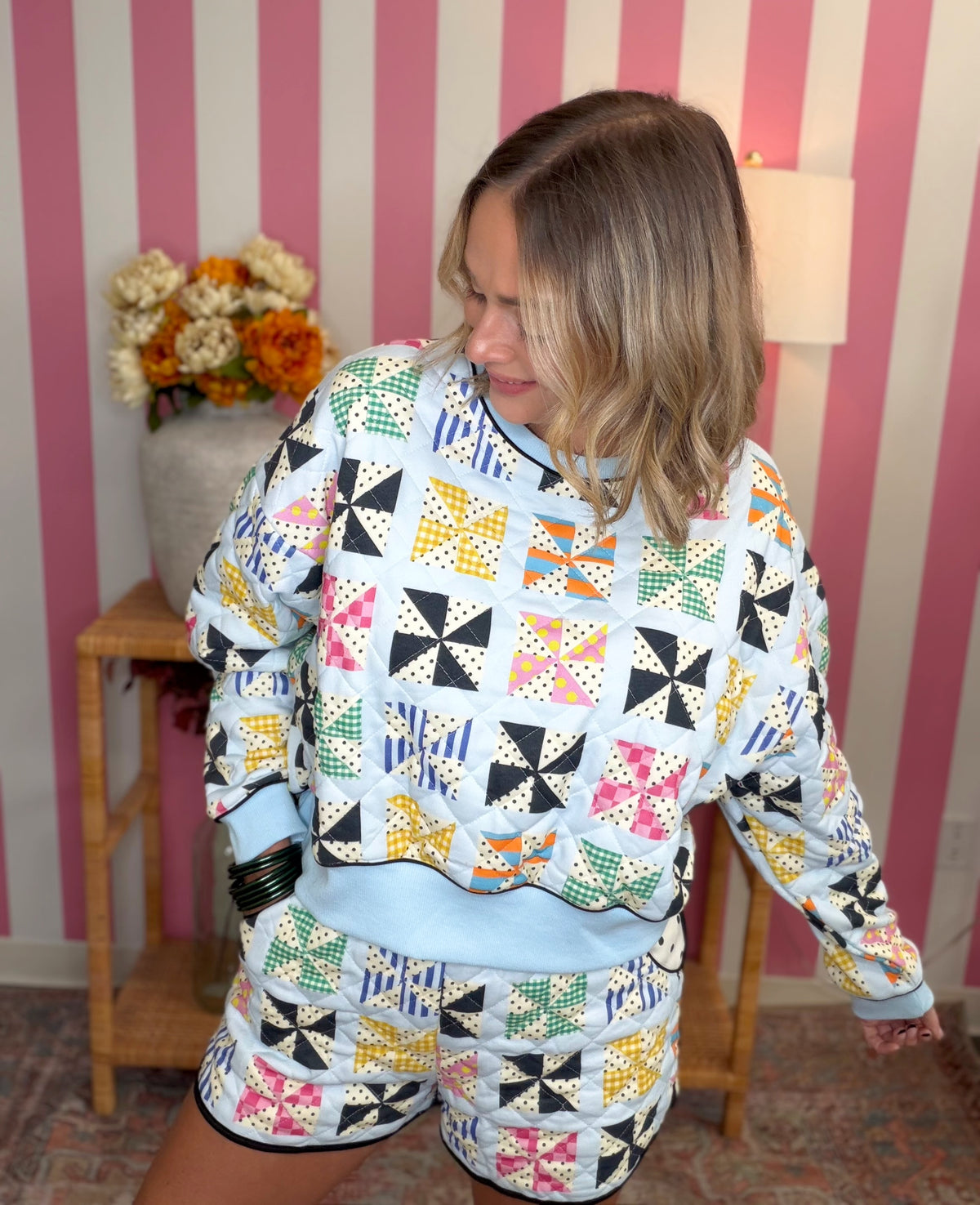 A woman wearing a matching two-piece lounge set with a colorful geometric patchwork print on a light blue base, standing in front of pink and white striped wallpaper.