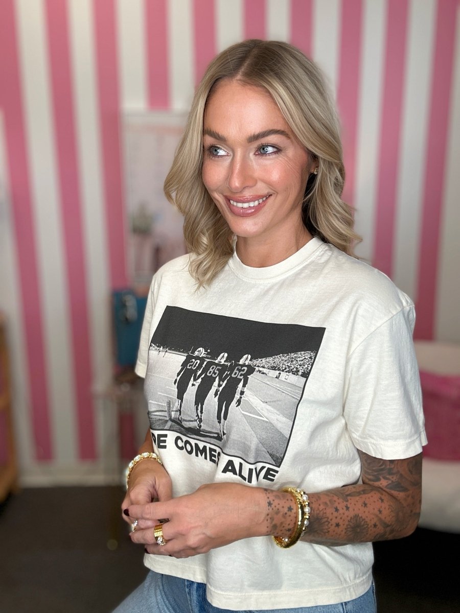 A woman with blonde hair wearing a white graphic t-shirt with a black and white band print that reads 'WE COME ALIVE'. She is standing in front of a pink and white striped wall.