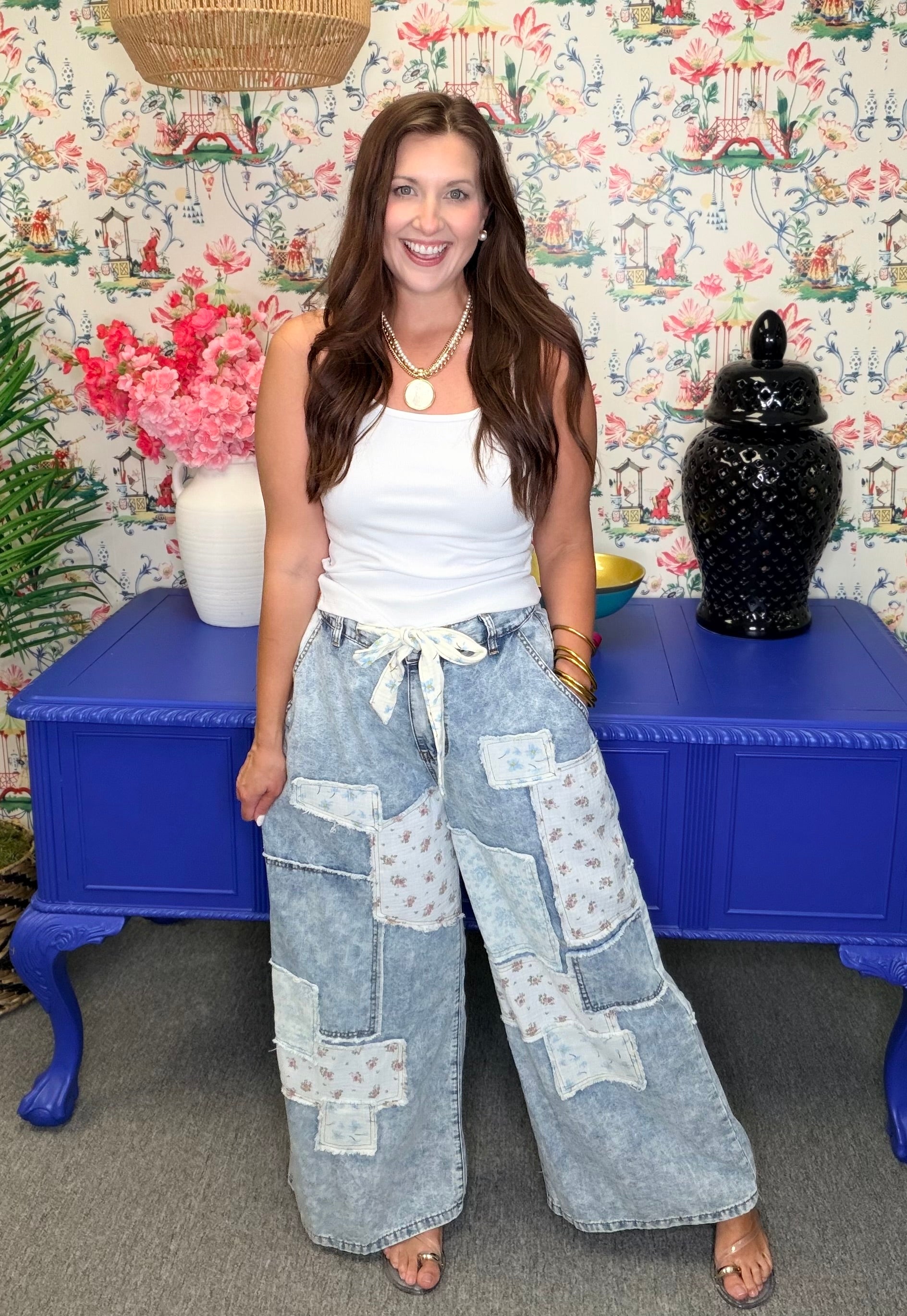 Woman wearing a white top and patchwork jeans standing in front of a colorful floral-patterned wall.
