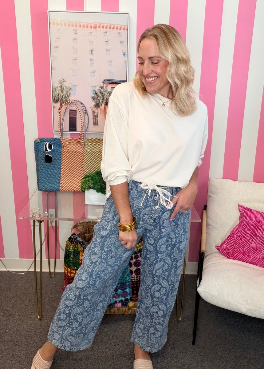 A woman posing in a white long-sleeve top and blue paisley wide-leg pants in a pink and white striped room.
