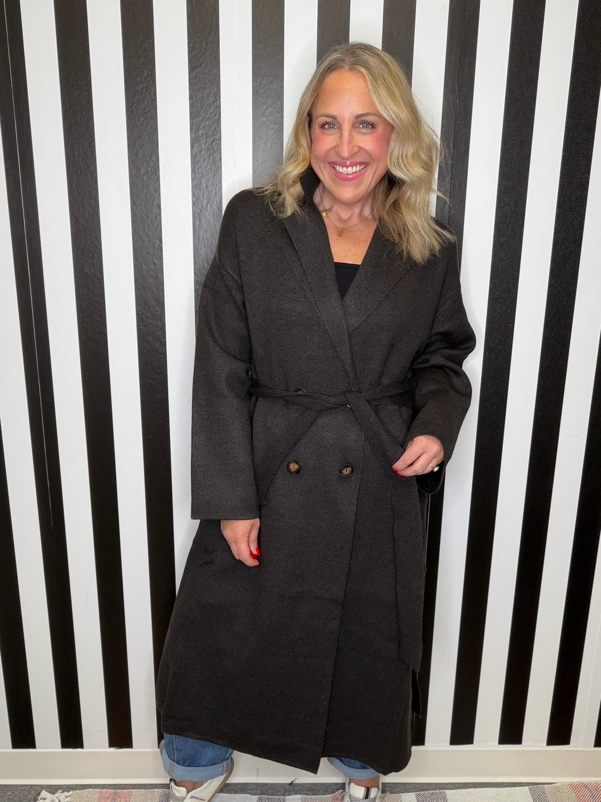 Woman in a brown trench coat standing in front of a black and white striped wall smiling.