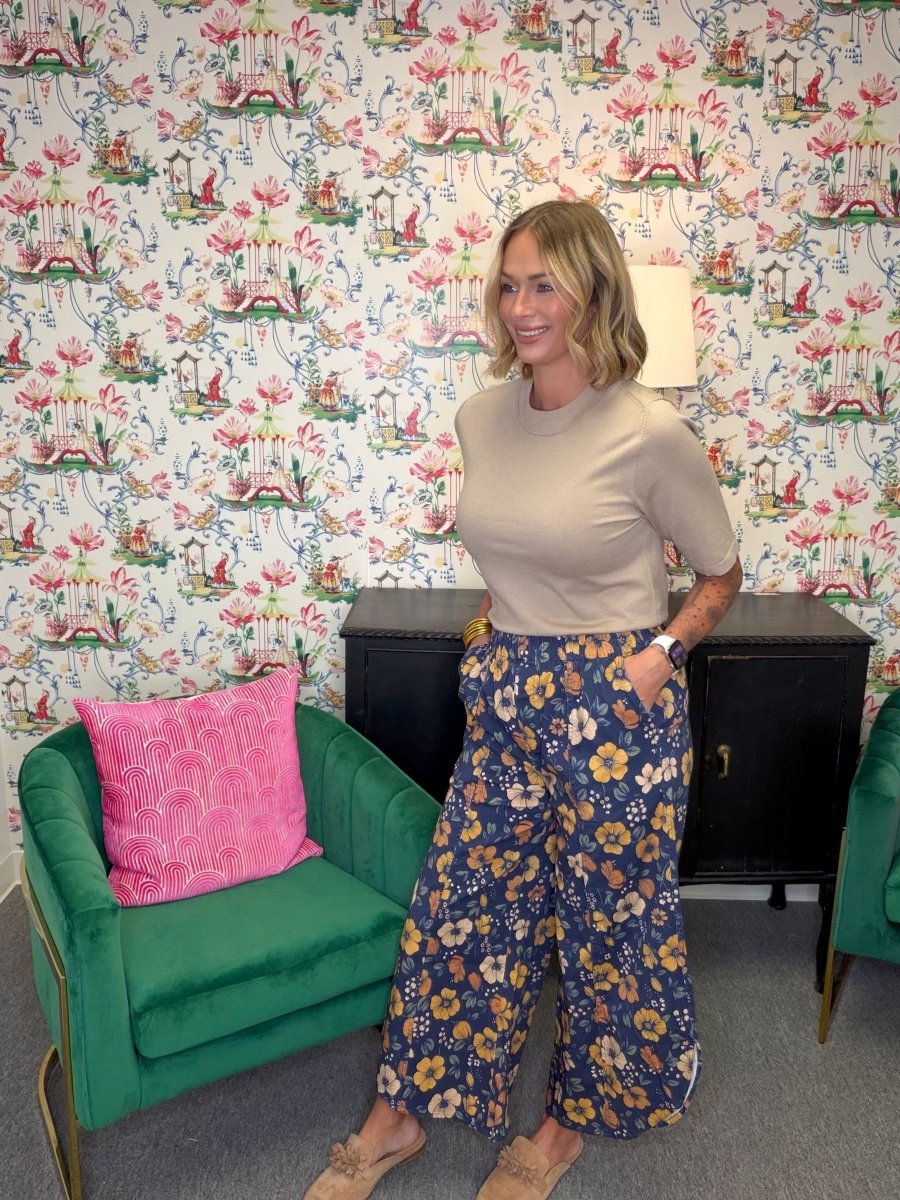 A woman stands in a room with floral wallpaper, wearing a beige short-sleeve top and navy wide-leg pants with a yellow and white floral pattern. She is standing next to a green velvet armchair with a pink pillow.