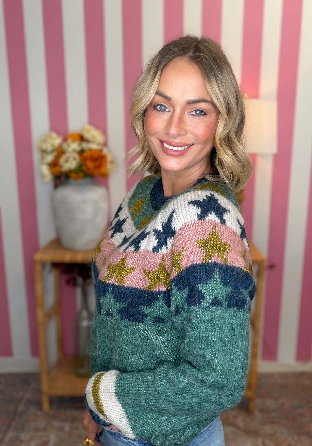 A woman wearing a colorful knit sweater with star patterns, standing in front of a pink and white striped wall with a wooden side table and flowers in the background.