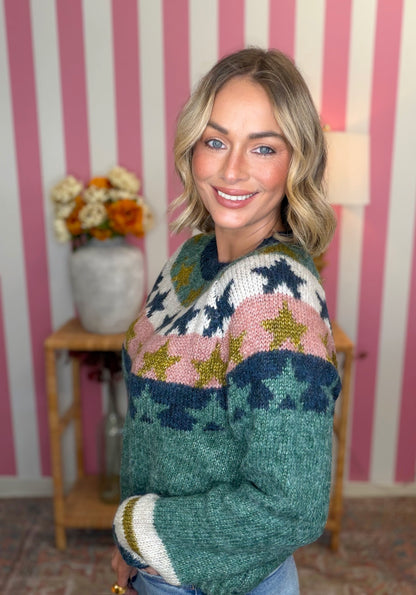 A woman wearing a colorful knit sweater with star patterns, standing in front of a pink and white striped wall with a wooden side table and flowers in the background.