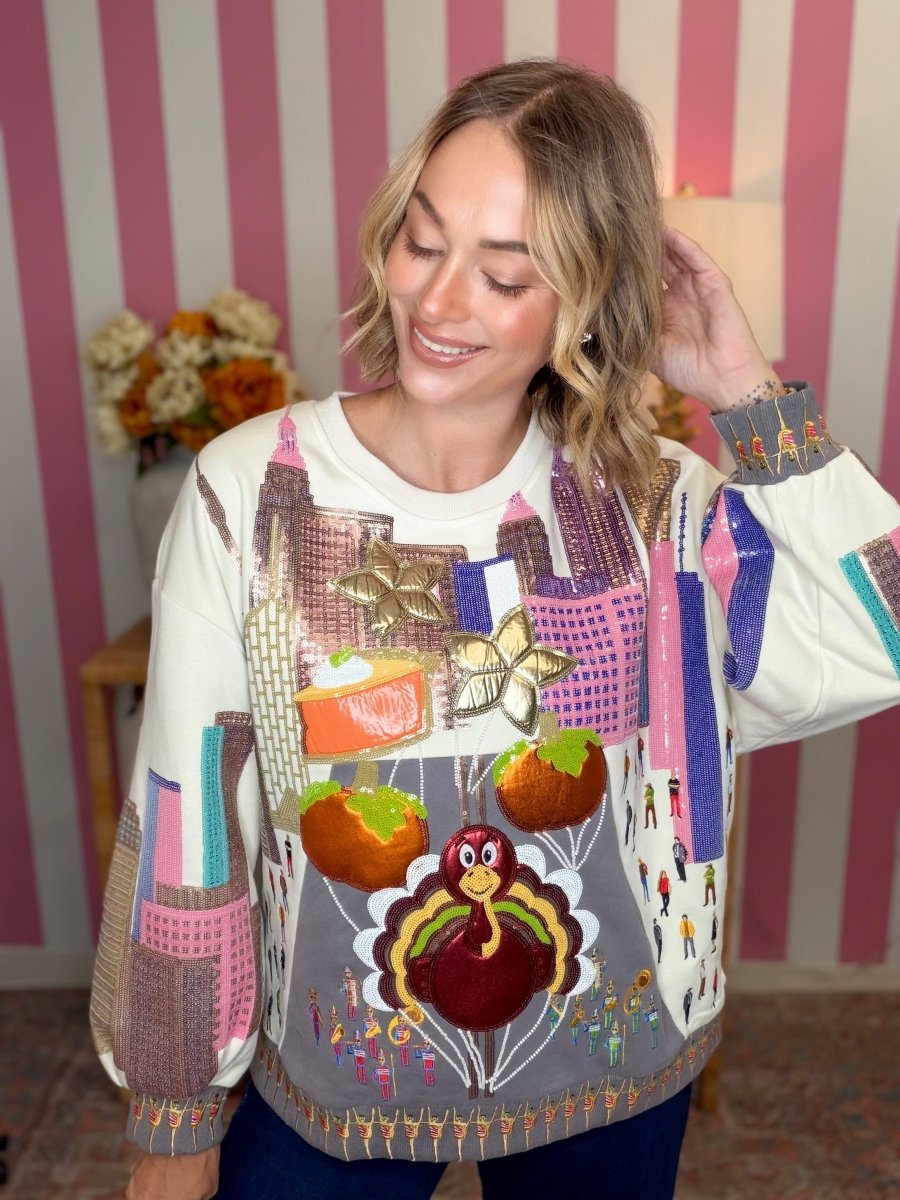 A woman wearing a long-sleeve crewneck sweater with a highly detailed, colorful Thanksgiving-themed patchwork design featuring a turkey, pumpkins, and a city skyline, posed against a pink and white striped wall.