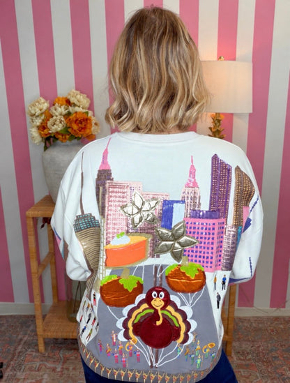 A person viewed from behind wearing a white crewneck sweatshirt with a highly detailed, colorful Thanksgiving-themed graphic print featuring a turkey, turkeys, pie, and a city skyline. The background features pink and white vertical stripes, a wooden side table with flowers, and a lamp.