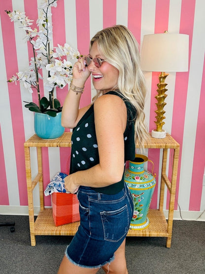 Woman in a polka dot top and denim shorts standing in front of a pink and white striped wall with decorative items.