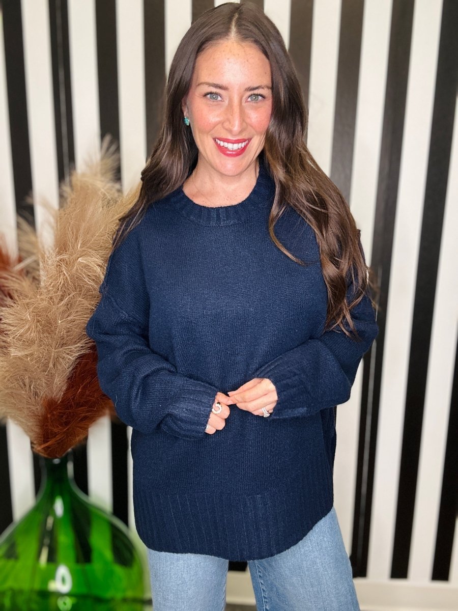 A woman with long brown hair smiling while wearing a dark navy blue knit crewneck sweater and light blue jeans, standing in front of a black and white striped wall.