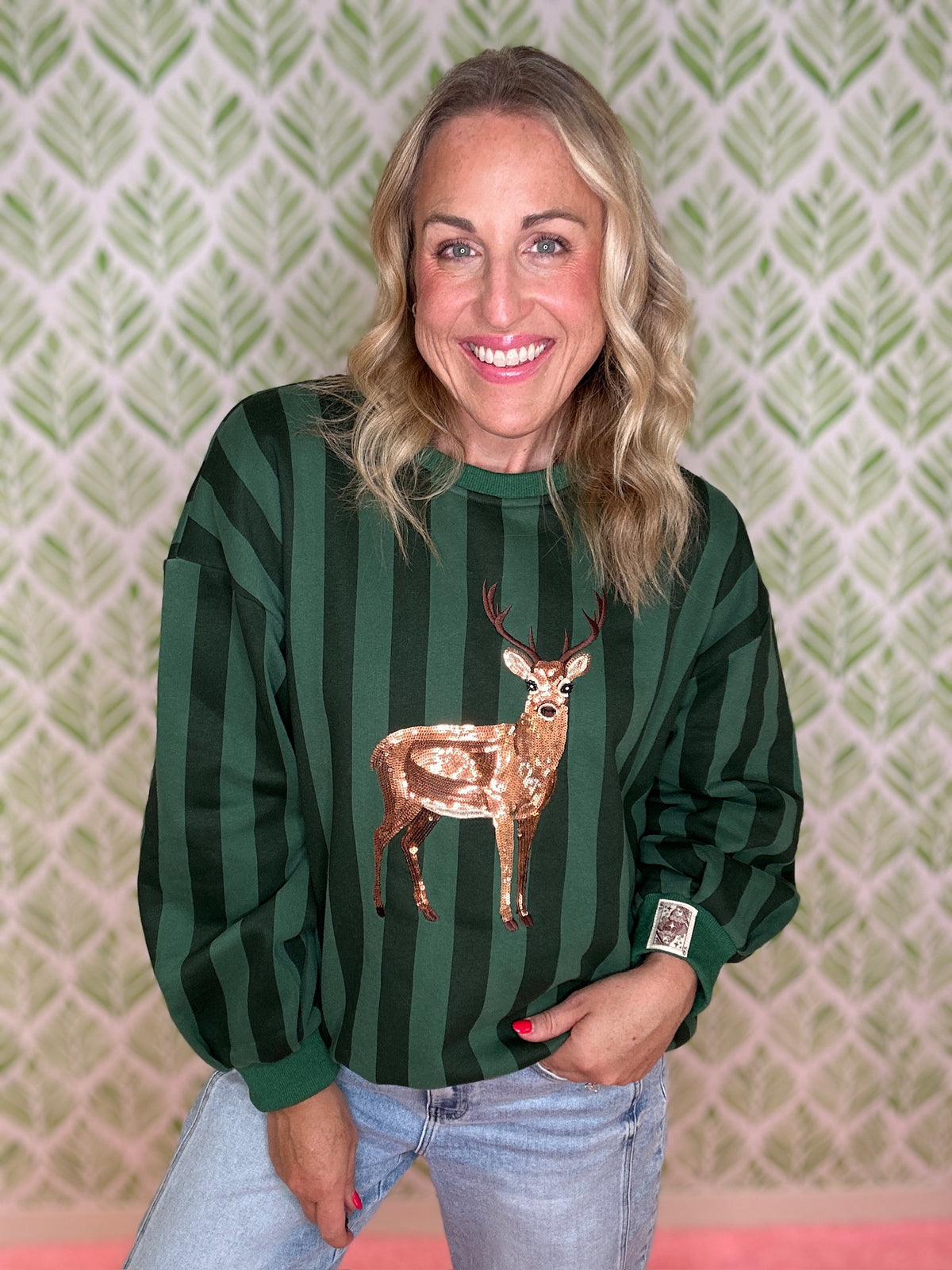 A smiling woman wearing a dark green and forest green vertically striped long-sleeve sweatshirt with a sequined deer applique on the front, paired with light blue jeans, standing against a green and white patterned wallpaper background.